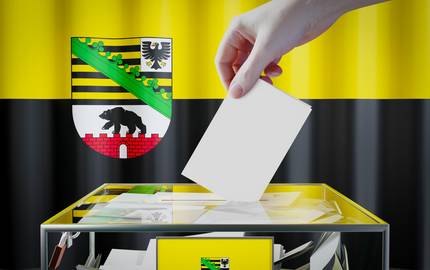 Eine Hand wirft einen Zettel in eine durchsichtige Wahlurne. Im Hintergrund die schwarz-gelbe Flagge und links oben das Wappen von Sachsen-Anhalt